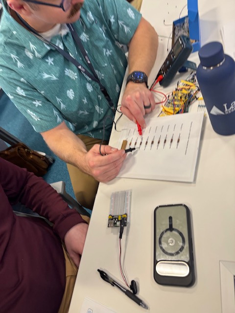 Hands use red and black test probes beside an electronics worksheet and a wired breadboard